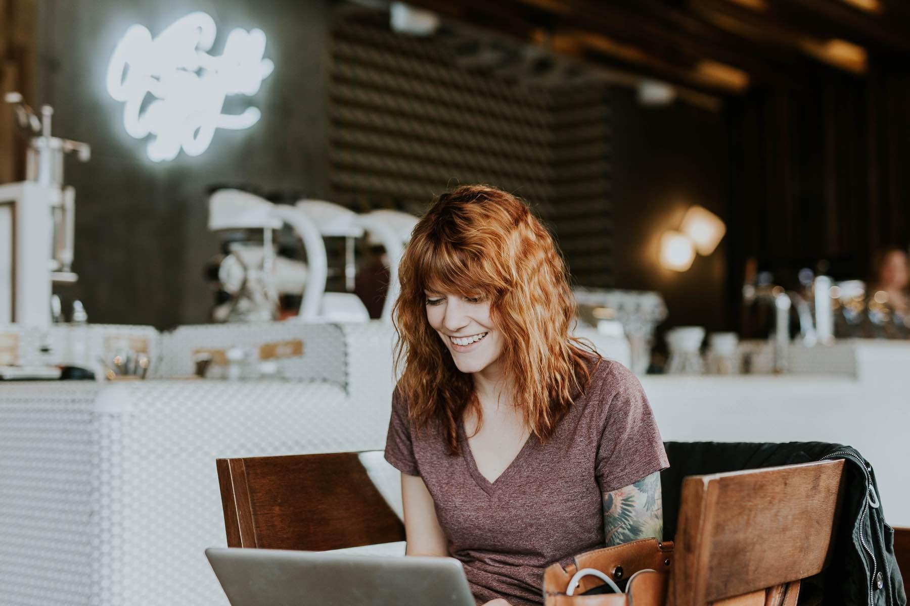 Mujer sonriente con cabello rojo utilizando portátil en un entorno de café.