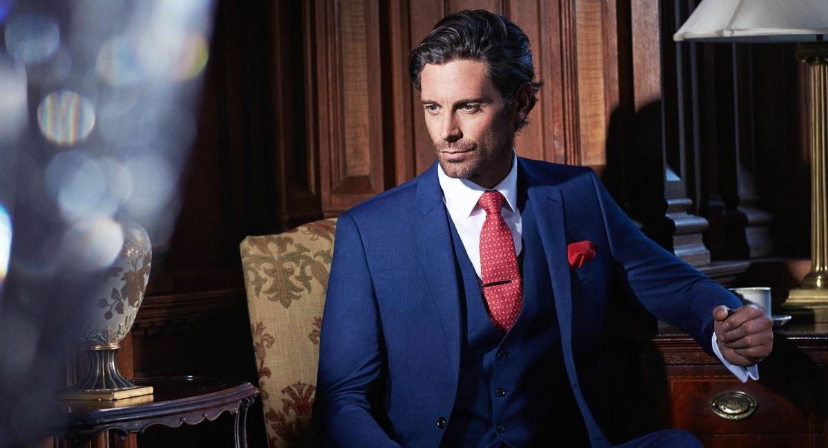 Man in elegant blue suit with red tie sitting by a wooden table.