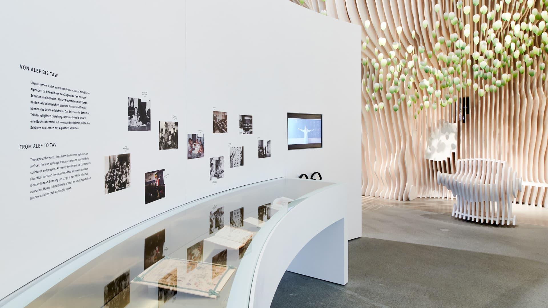 Innenansicht eines modernen Ausstellungsraums mit Texttafeln, Fotografien und interaktiven Displays.