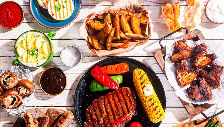 Nourriture grillée variée comprenant des côtes, du maïs et des pommes de terre sur une table en bois.