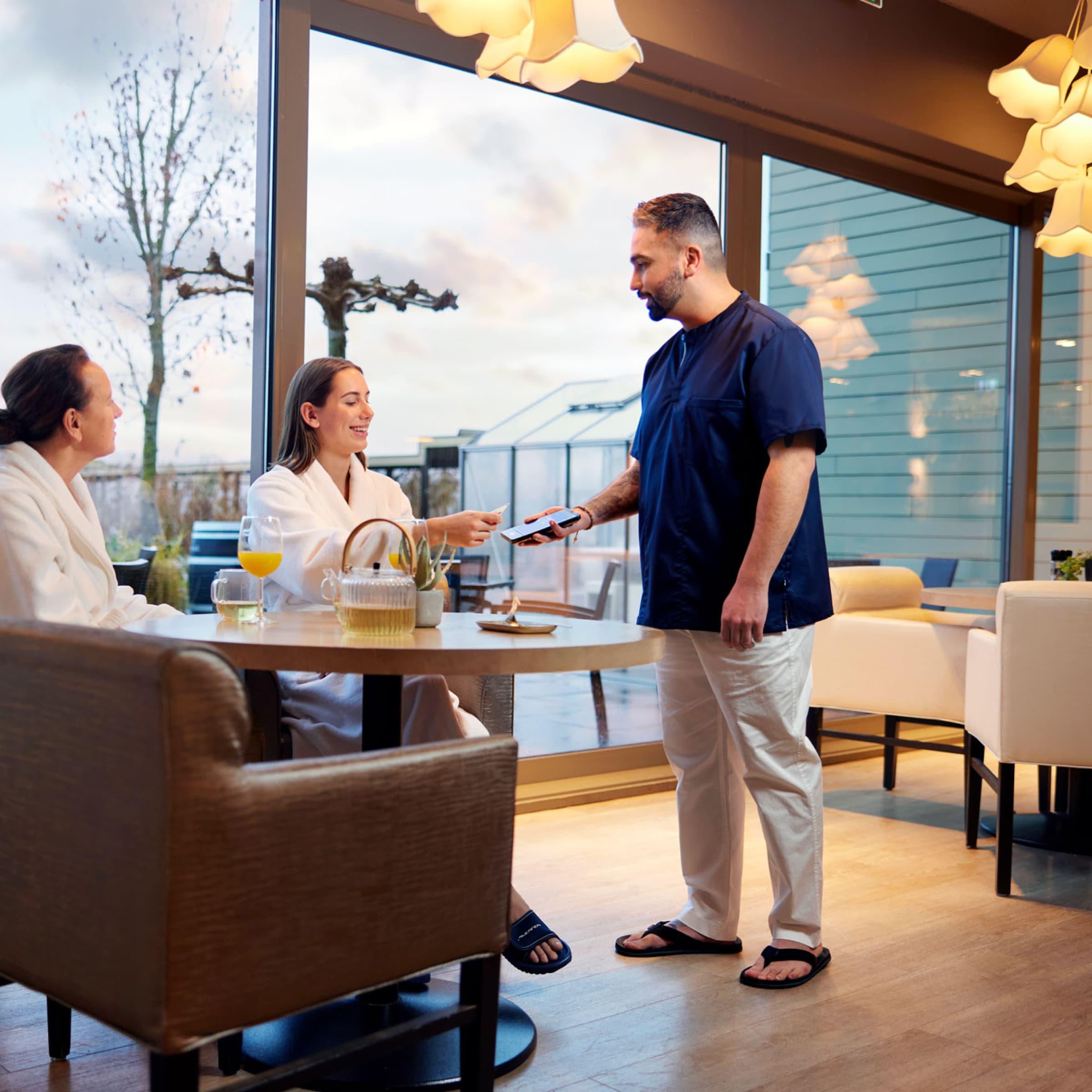 Waiter using a mobile payment device to accept a payment from a seated female customer at a restaurant.