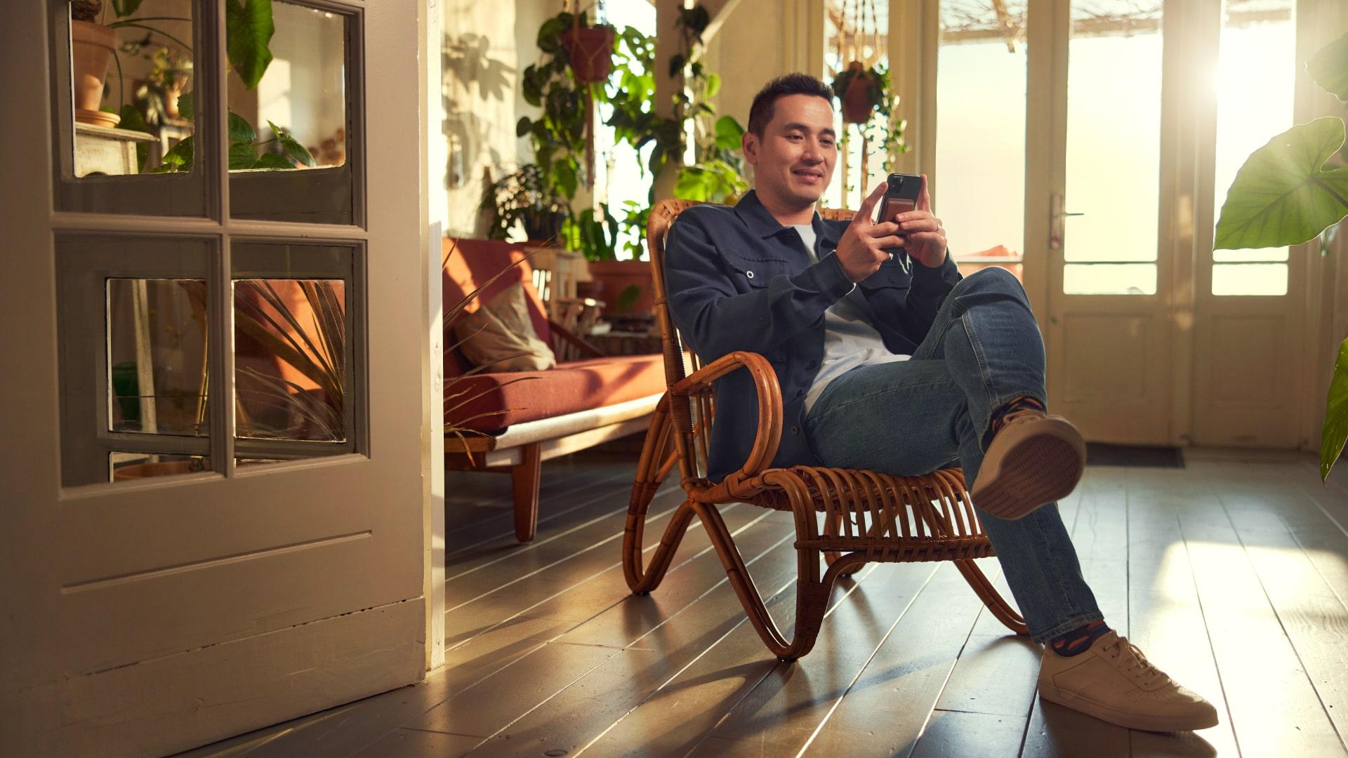 Hombre sentado en una mecedora de rattán utilizando un smartphone en una habitación soleada con plantas.