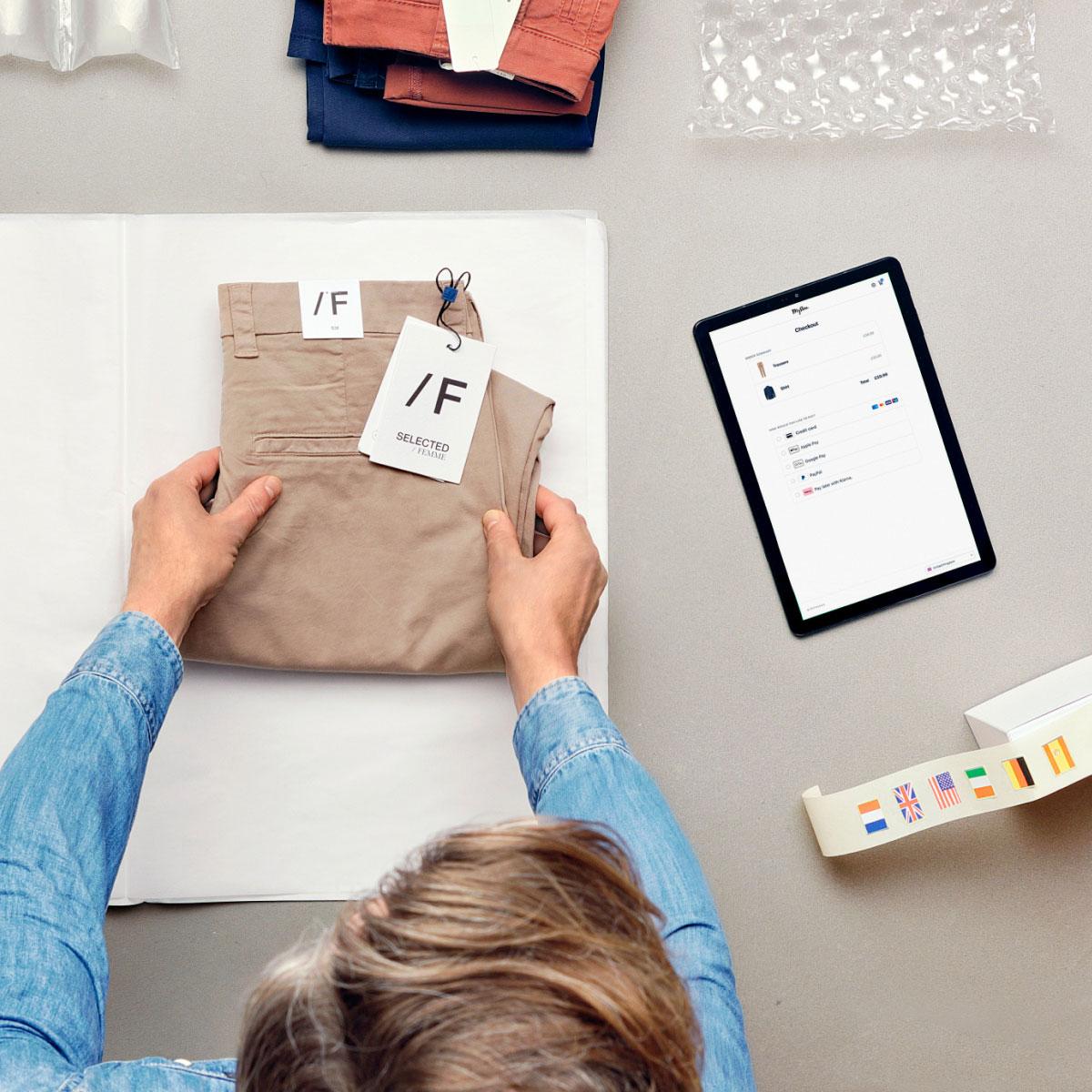 Persona empaquetando un par de pantalones beige con etiqueta, junto a una tableta que muestra la interfaz de pago de Adyen.