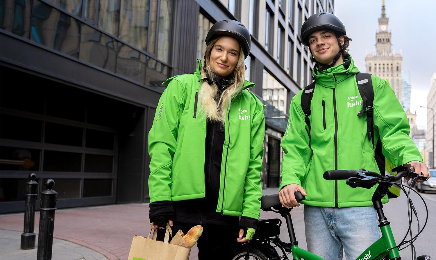 Dwie osoby dostarczające w jasnozielonych kurtkach trzymające papierową torbę z widokiem na miasto w tle.