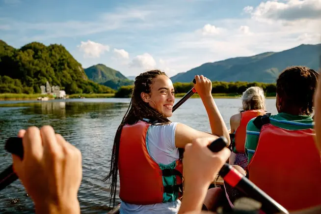 Menschen rudern ein Boot mit Rettungswesten an einem sonnigen Tag mit Bergen im Hintergrund.