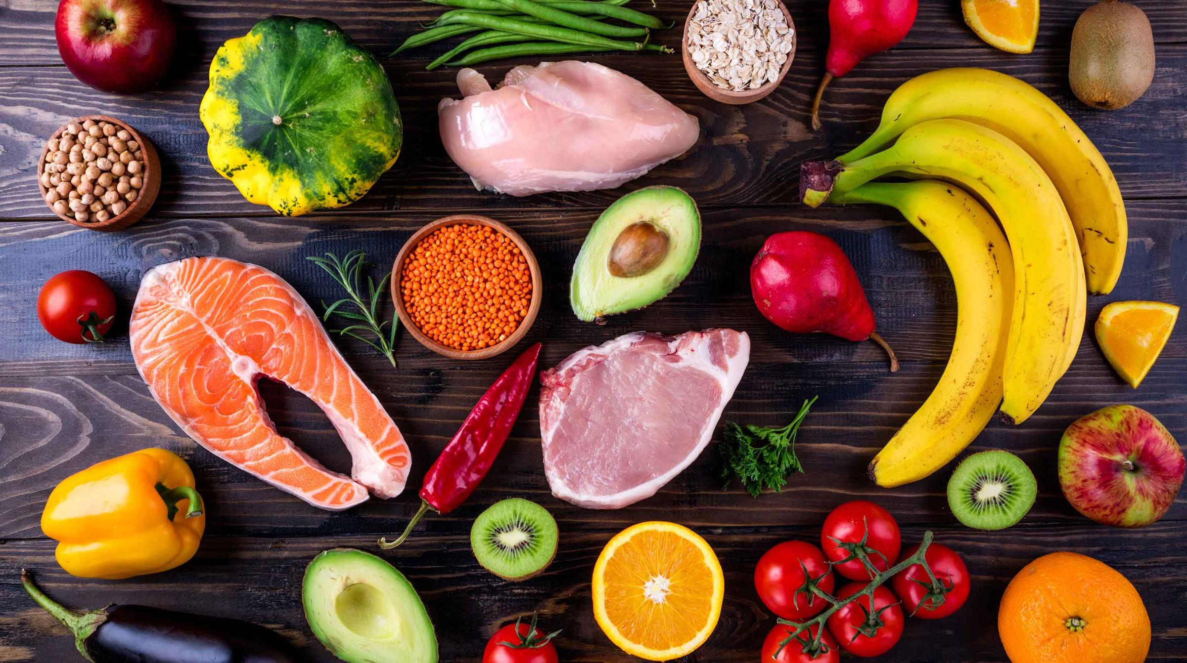 Assortment of healthy foods including fruits, vegetables, and meats arranged on a wooden surface.