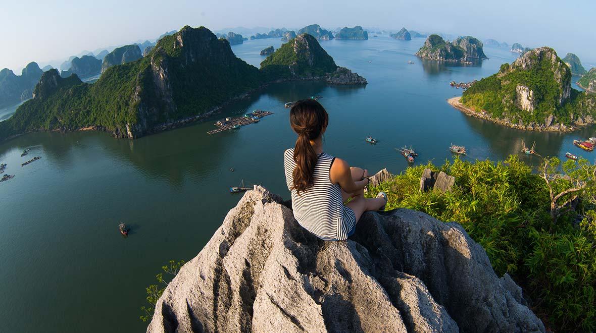 Person sitzt auf einem Berggipfel mit Blick auf eine malerische Bucht mit Booten und Inseln