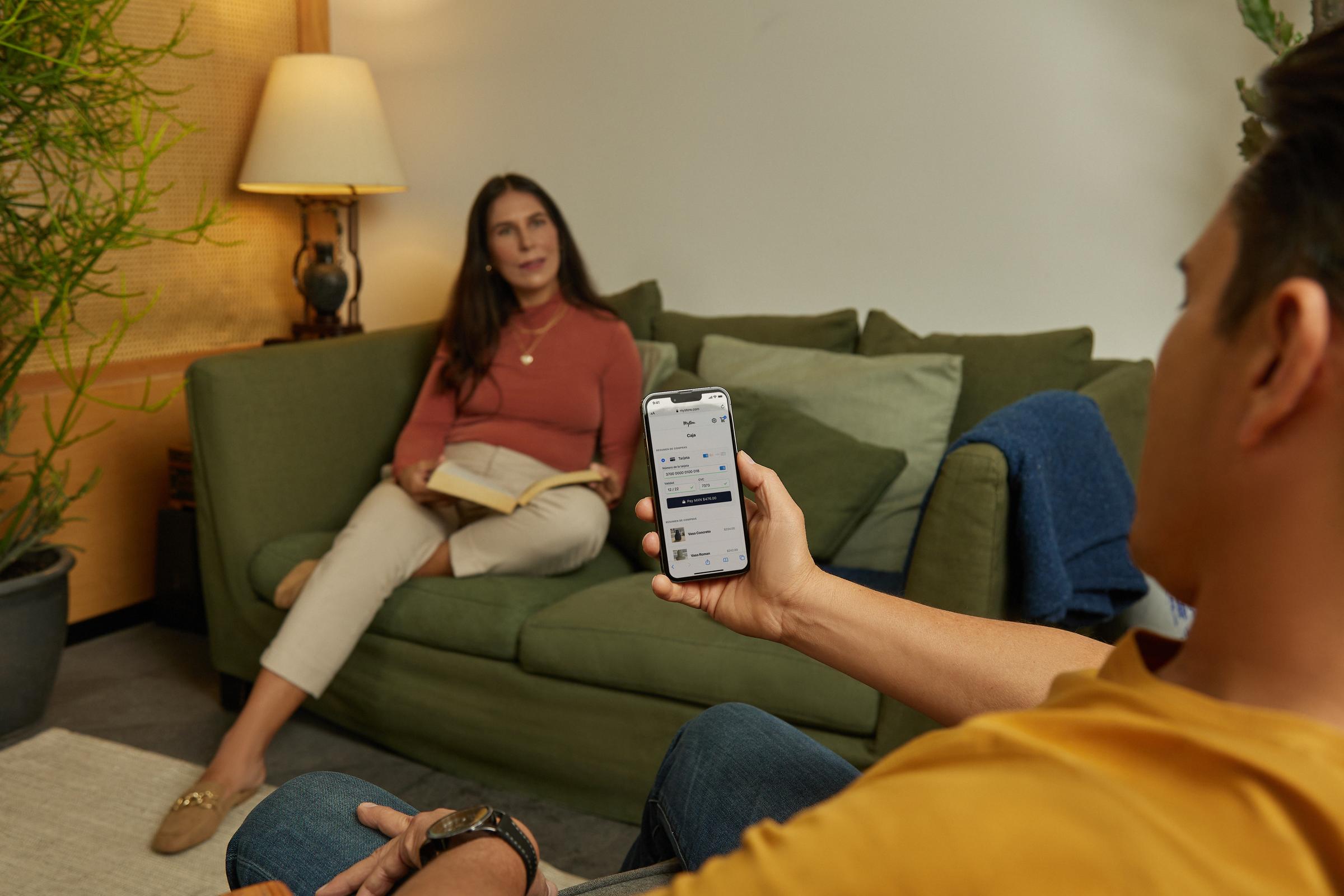 Persona que sostiene un smartphone mostrando la interfaz de pago de Adyen, otra persona en el fondo en el sofá.