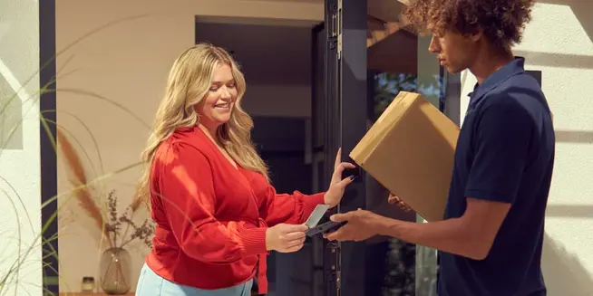 Persona che consegna un pacco e un lettore di carte a una donna sorridente sulla sua porta di casa.