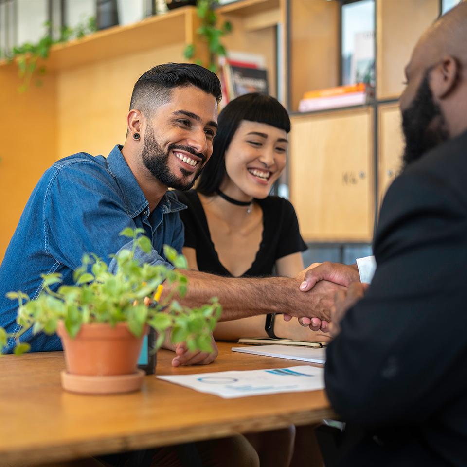 Two people shaking hands across a table with paperwork, another person observing with a smile.