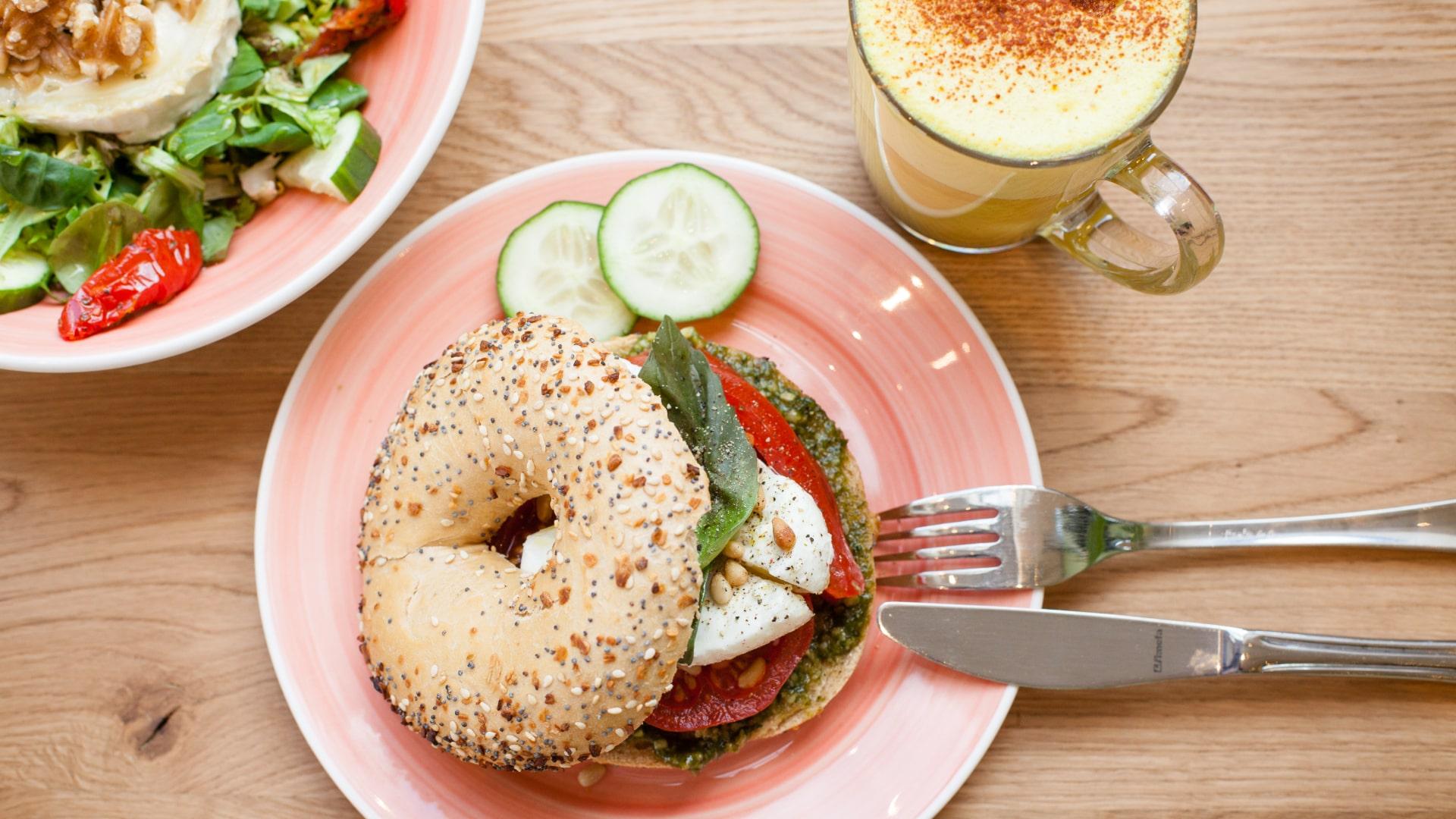 Allt-i-ett-bagel med cream cheese, spenat, tomat och gurka på en rosa tallrik med en sallad och en kryddlat latte.