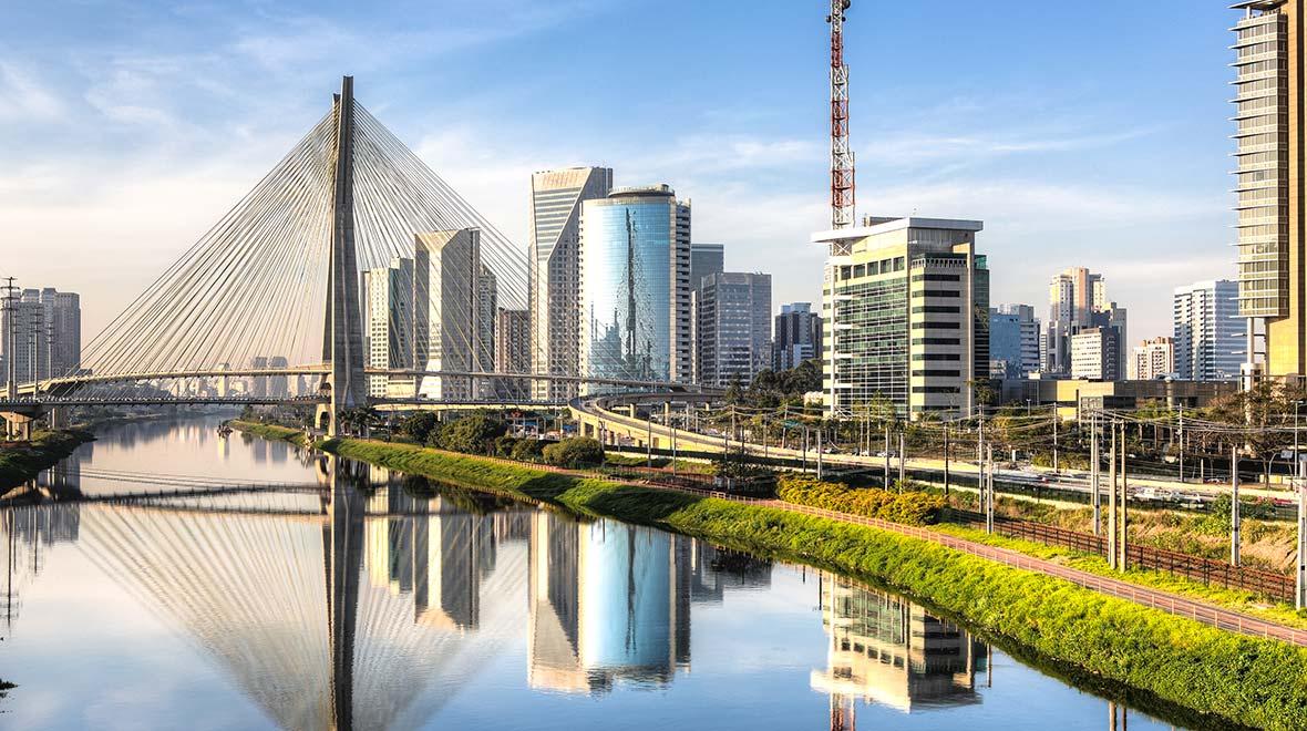 Cenário urbano moderno com uma ponte estaiada sobre um rio tranquilo refletindo os edifícios ao redor.