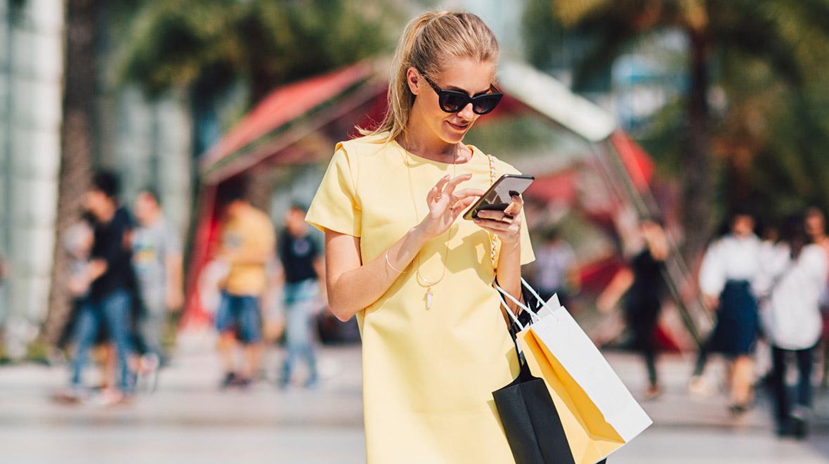 Frau in gelbem Kleid und Sonnenbrille nutzt Smartphone, während sie Einkaufstüten trägt.