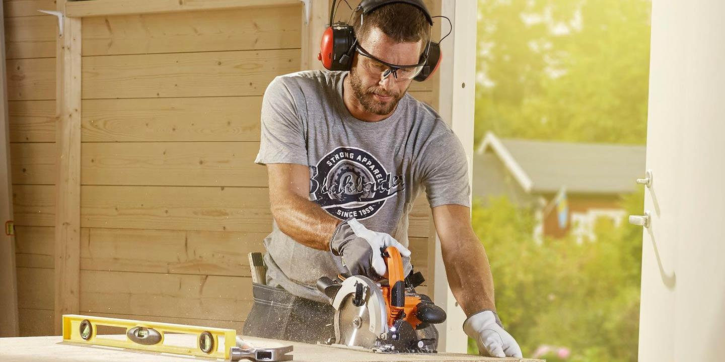 Man använder en cirkelsåg på en träplanka i en solig utomhusverkstad med säkerhetsutrustning.