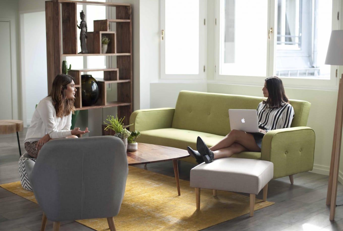 Zwei Frauen in einem modernen Wohnzimmer, eine auf einer limettengrünen Couch mit Laptop, die andere auf einem grauen Stuhl.