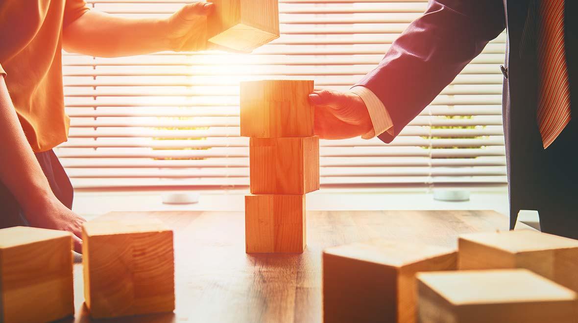 Duas pessoas constroem uma torre com blocos de madeira em uma mesa com luz solar ao fundo.
