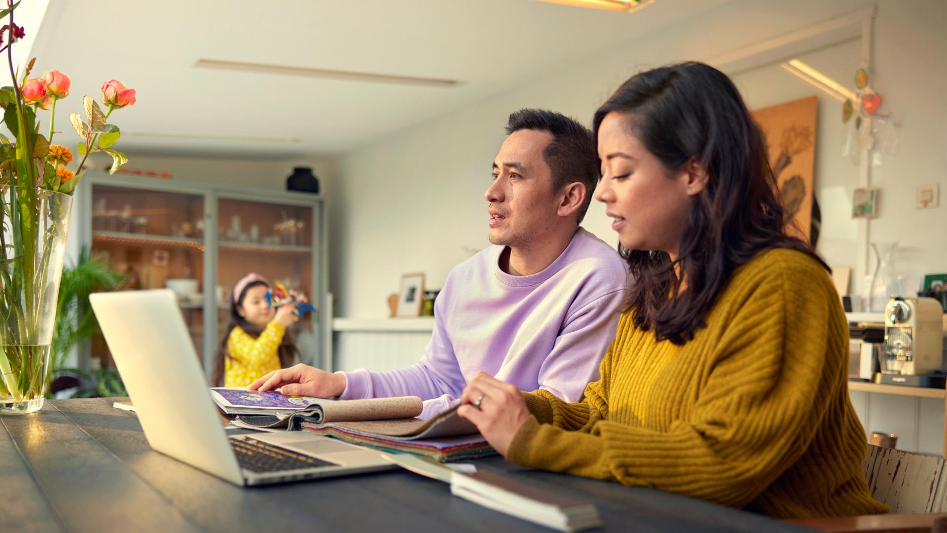 Deux adultes regardant un ordinateur portable tandis qu'un enfant joue en arrière-plan.