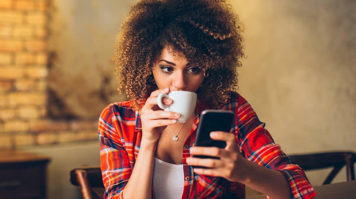 Mulher de cabelos cacheados tomando café e olhando para o smartphone.