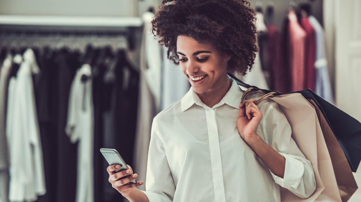 Mulher sorrindo com sacolas de compras usando smartphone na loja.
