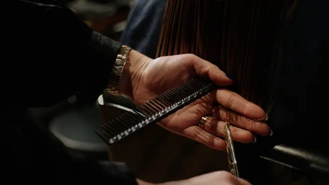 Duas mãos seguram um pente e tesouras enquanto cortam um fio de cabelo