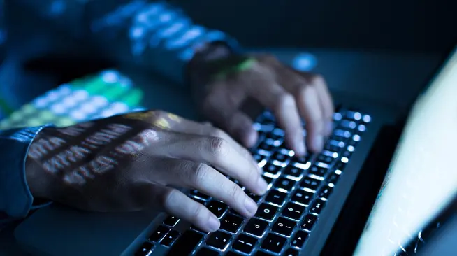 Close-up of hands typing on a backlit laptop keyboard with screen reflection.