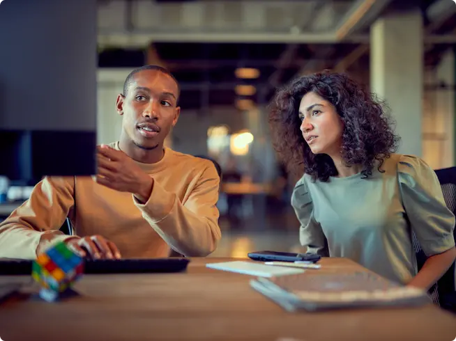 Dos colegas discutiendo trabajo con una computadora y un cubo de Rubik en el escritorio.