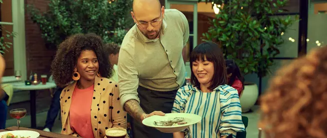 A group of people enjoying an outdoor meal with a server offering dishes.
