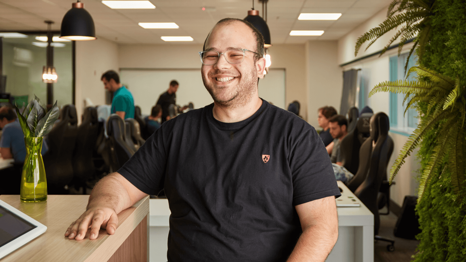 Homem sorridente com óculos e camiseta preta sentado em um ambiente de escritório