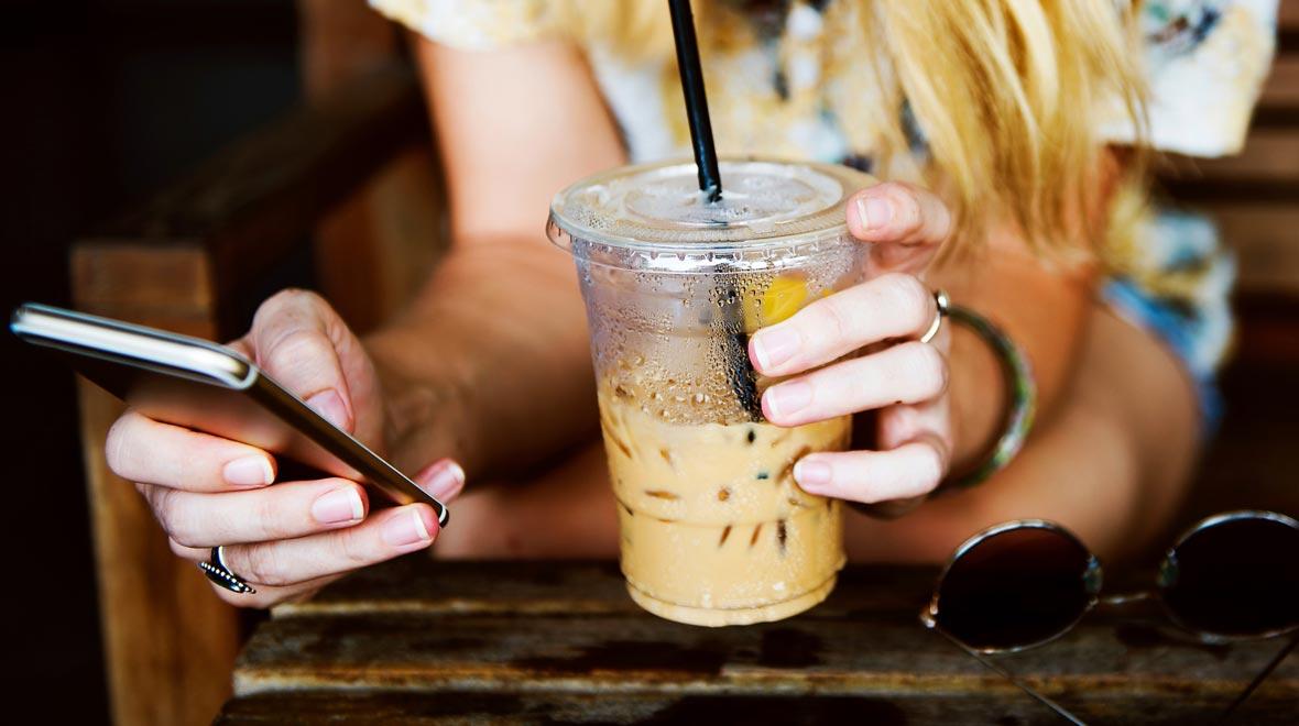 Primer plano de una persona que sostiene un smartphone en una mano y un café con hielo en la otra.