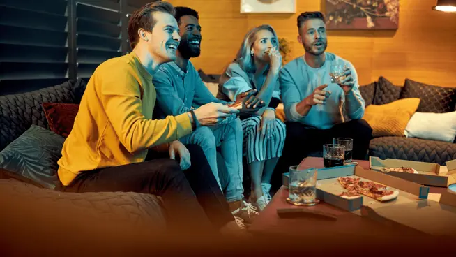 Four friends excitedly playing video games on a couch with snacks and drinks.