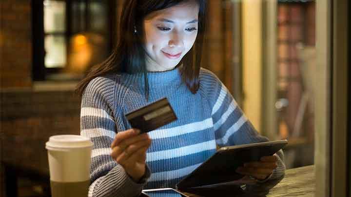 Mujer utilizando una tableta y sosteniendo una tarjeta de crédito con una taza de café en la mesa.