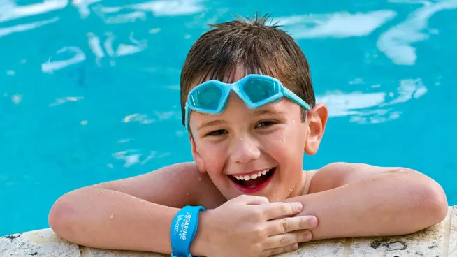 Smiling young boy with goggles resting by a swimming pool, wearing a blue Adyen wristband.