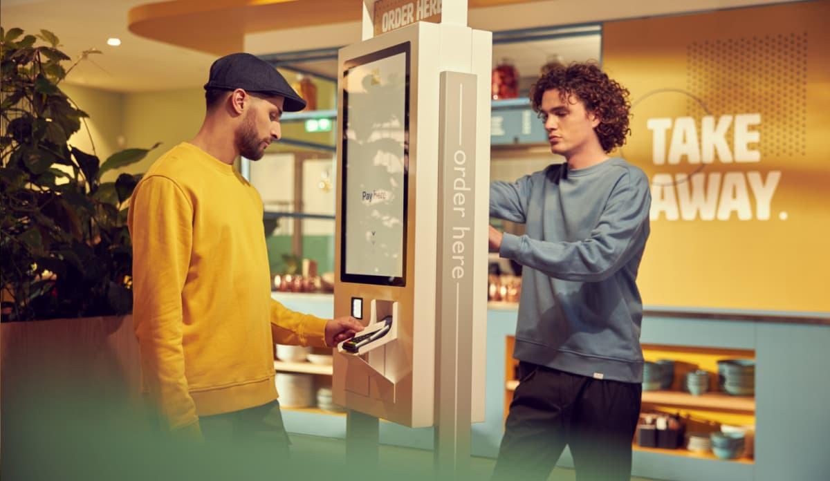 Duas pessoas em um quiosque de autoatendimento com terminal de pagamento sem contato Adyen em um café moderno.