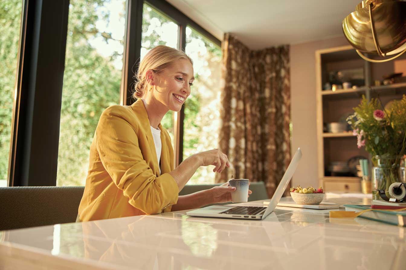 Mulher sorrindo em blazer amarelo usando laptop e segurando uma xícara de café em casa