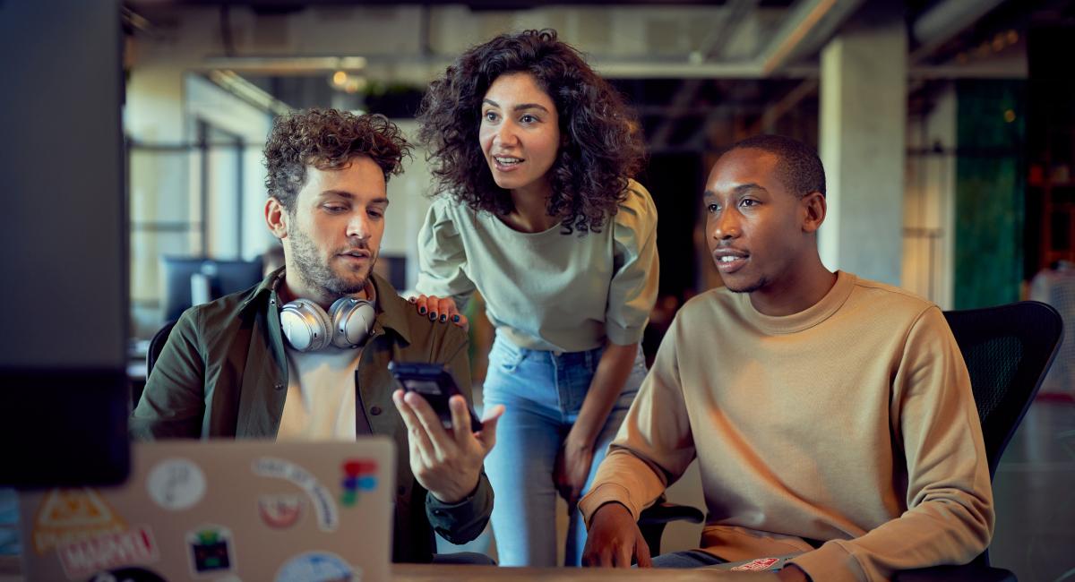Three colleagues collaborating with one showing a mobile screen to the others in a modern office setting.