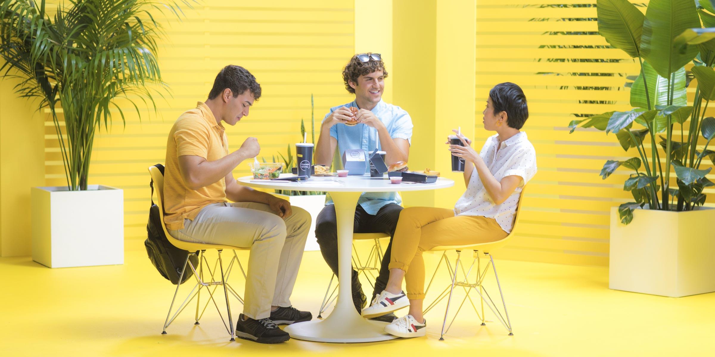 Three people enjoying a meal and interacting at a white table in a brightly colored yellow room.