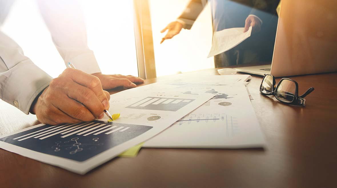 Persona analizando gráficos financieros y tomando notas con un colega en el fondo.