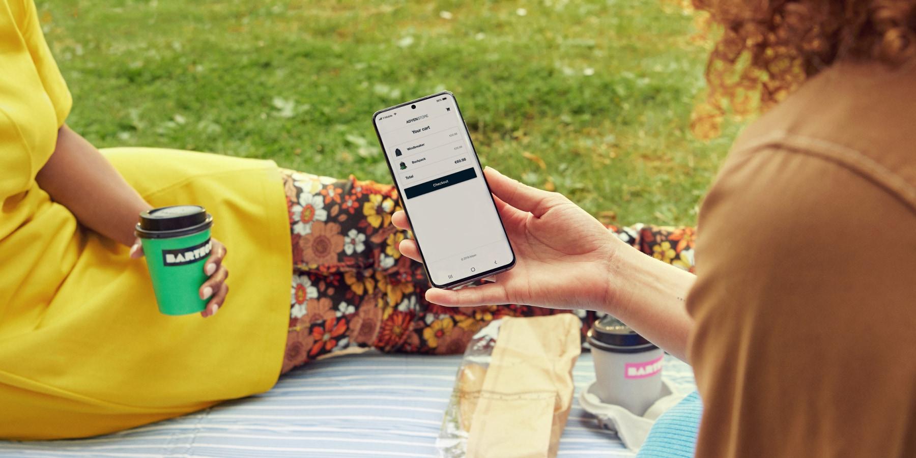 Person holder en smartphone viser en indkøbskurv på skærmen, med en takeaway kaffe og en sandwich i nærheden.