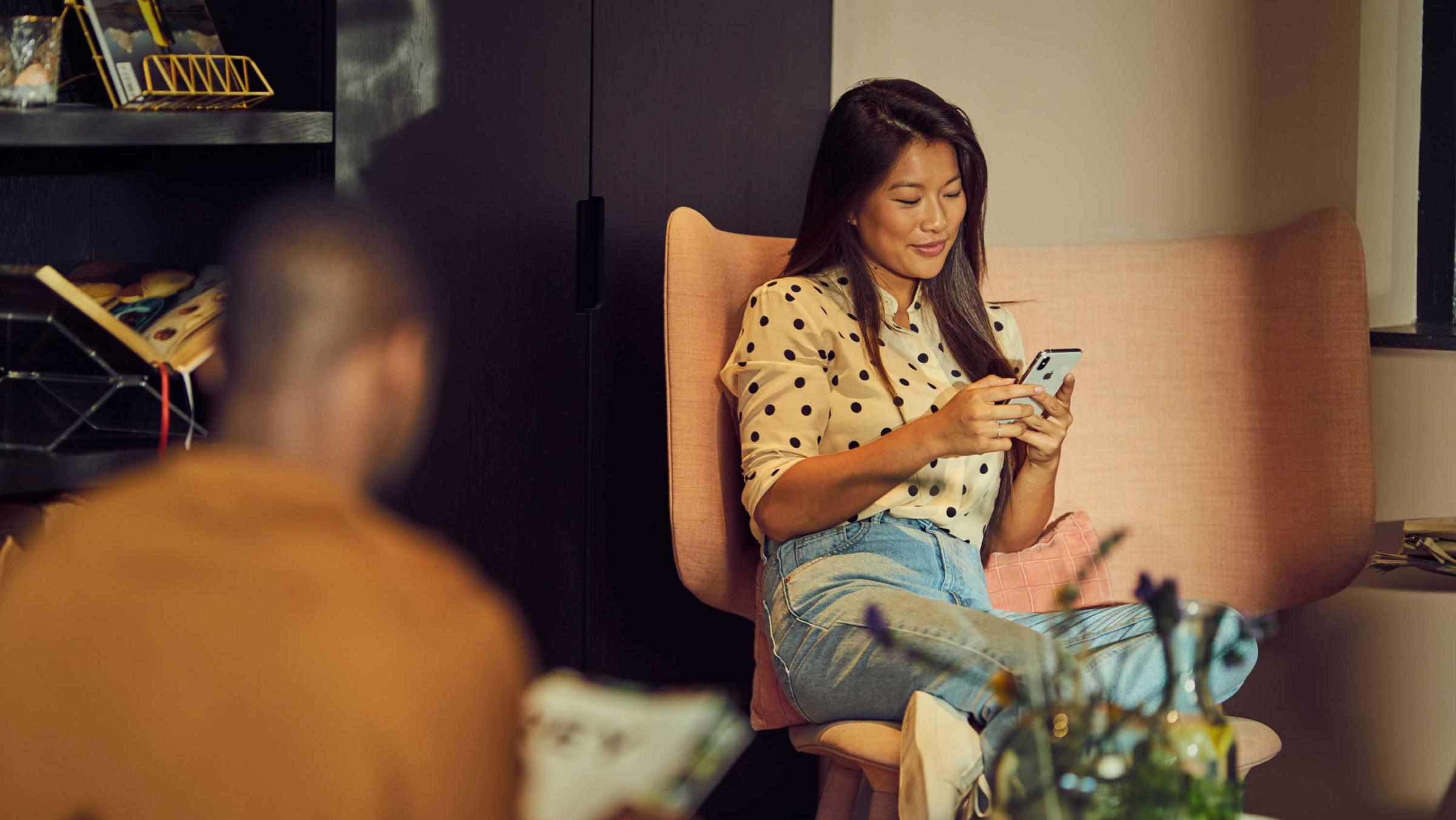 Frau sitzt in einem rosa Sessel und nutzt ein Smartphone, während ein Mann im Vordergrund unscharf ist.