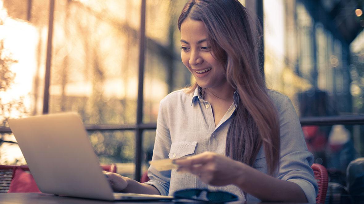 Mujer sonriendo usando portátil y sosteniendo tarjeta de crédito en terraza de café al aire libre.
