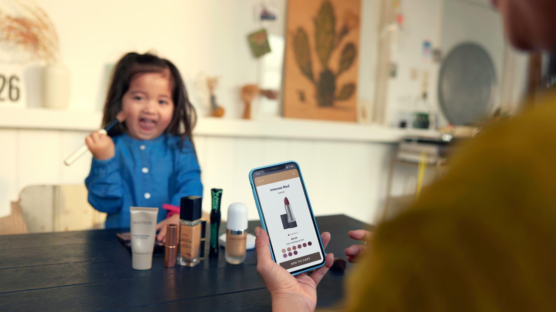 Person holding a smartphone with a cosmetics shopping app open, with a child in the background.