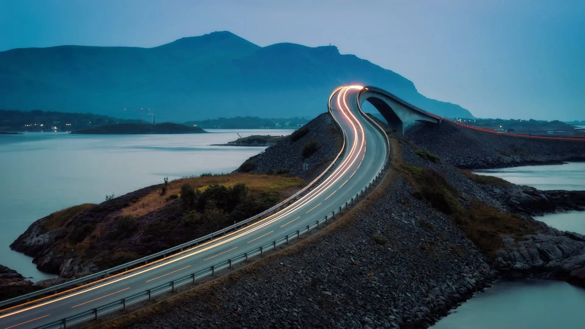 Curved bridge over a coastal inlet at dusk with light trails from passing vehicles in Norway.
