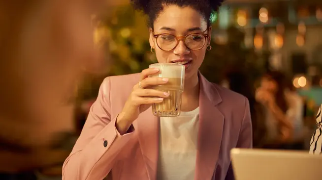 Woman wearing glasses, drinking a latte at a cafe with a laptop on the table