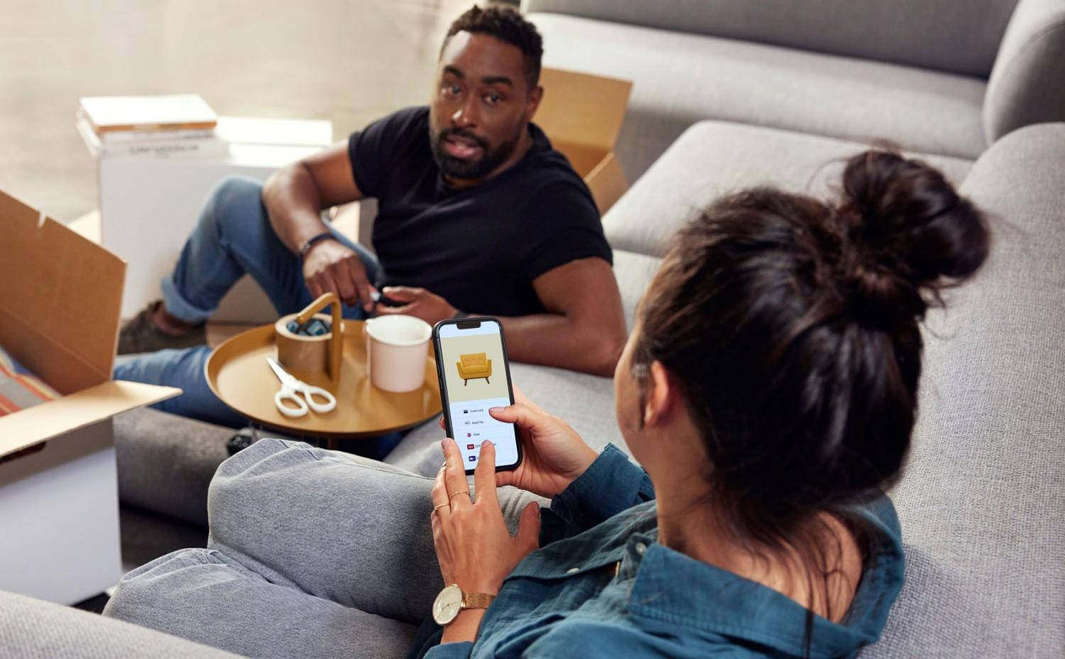 Two people in a living room, one holding a phone with a furniture shopping app open.