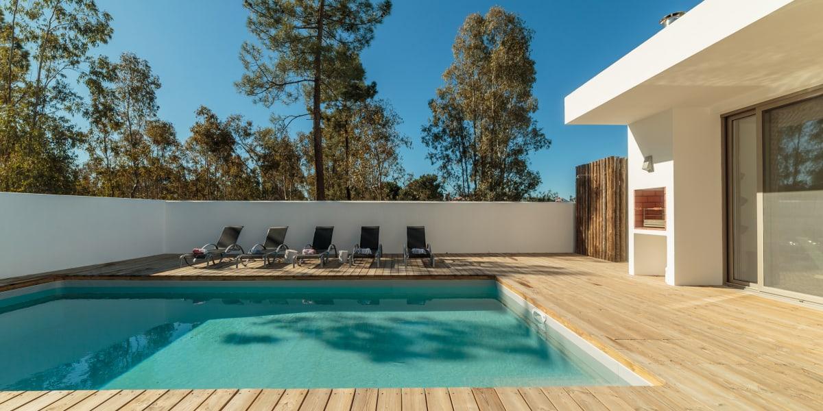 Maison moderne avec une terrasse en bois au bord de la piscine et des transats entourés d'arbres.