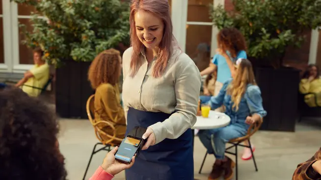 Persona sonriendo que acepta un pago móvil en un café al aire libre soleado.