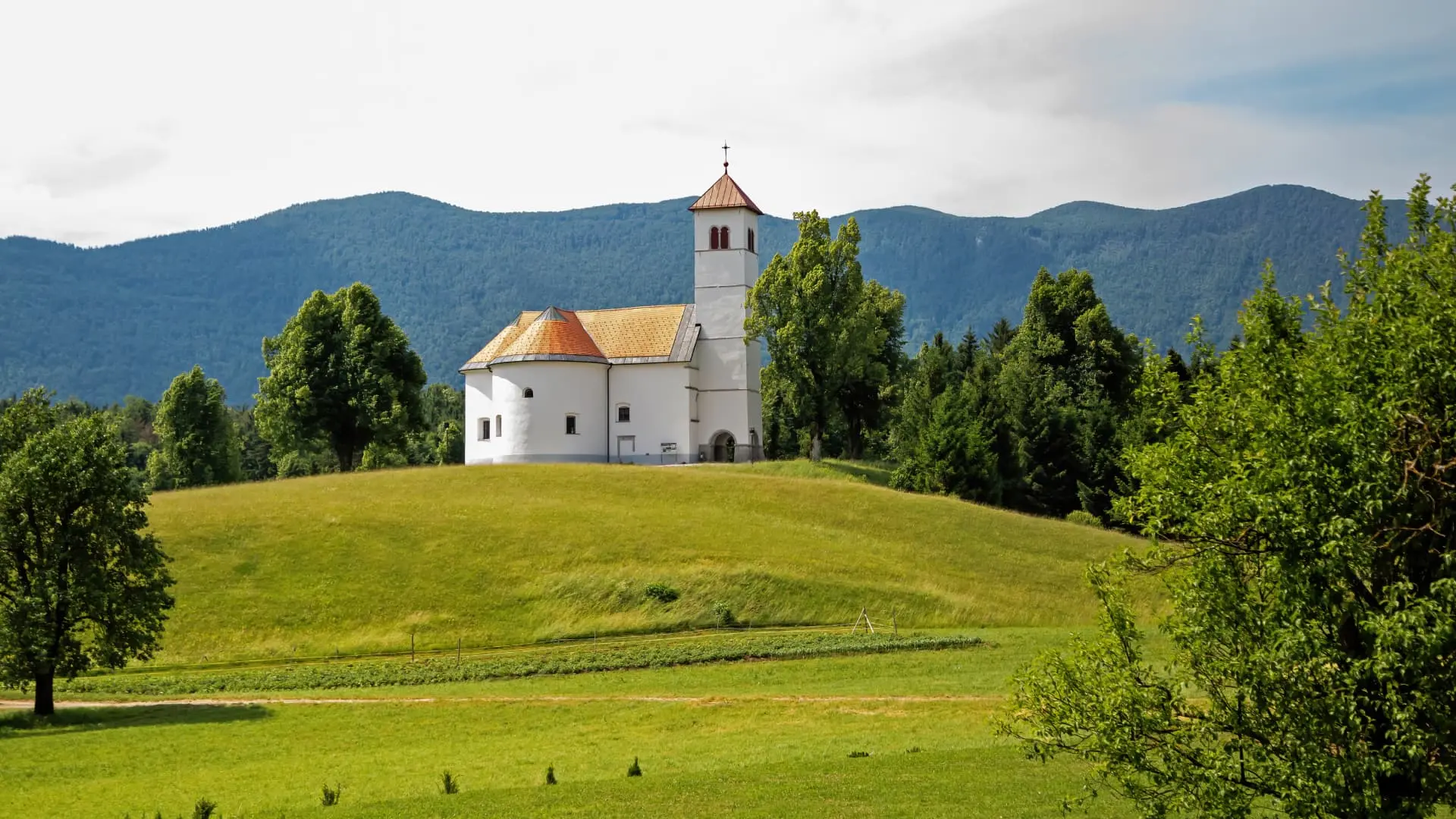 White church with a red roof on a green hill against a backdrop of mountains and trees in Slovenia.