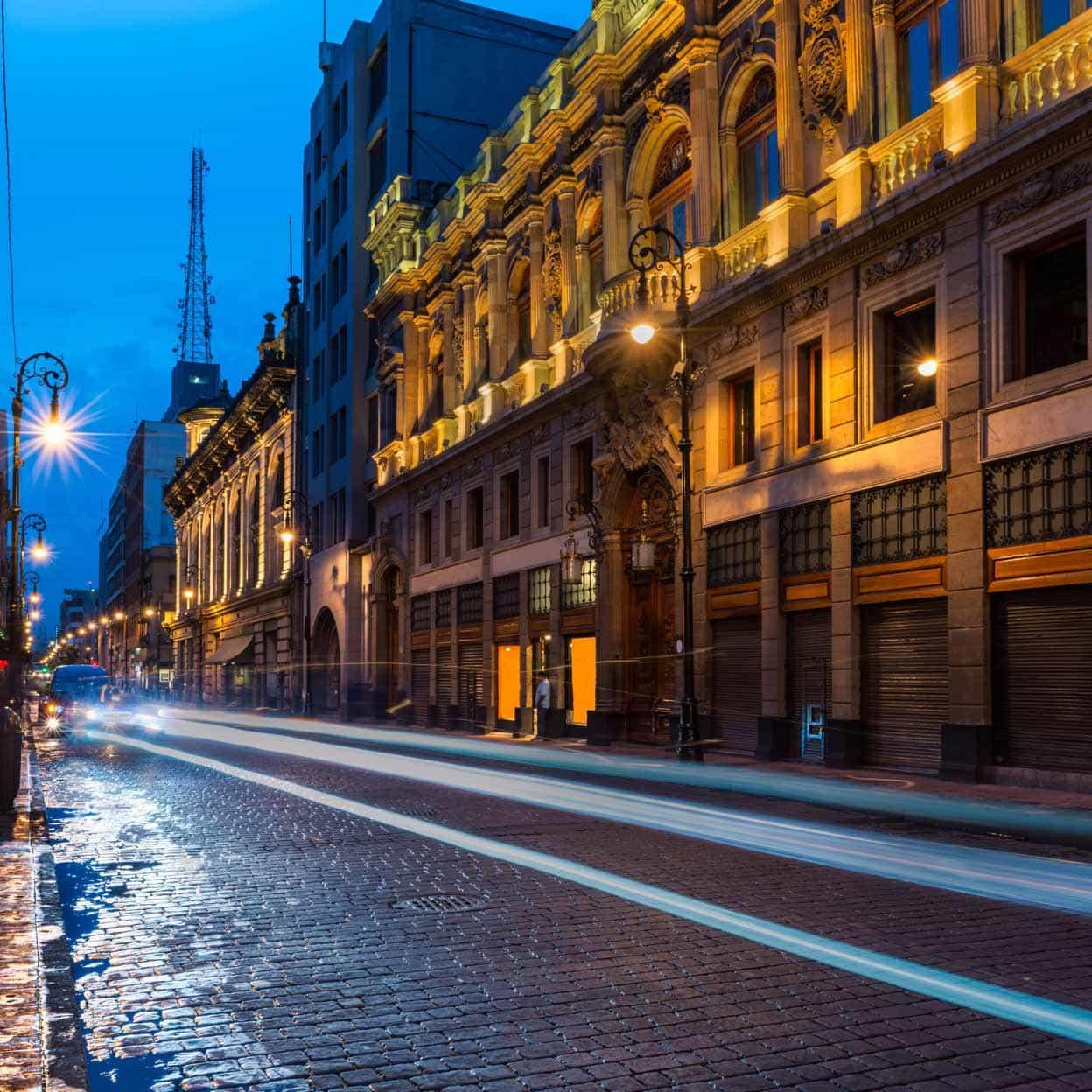 Vue de la ville au crépuscule avec des bâtiments éclairés et des traces de lumière de véhicules en mouvement.