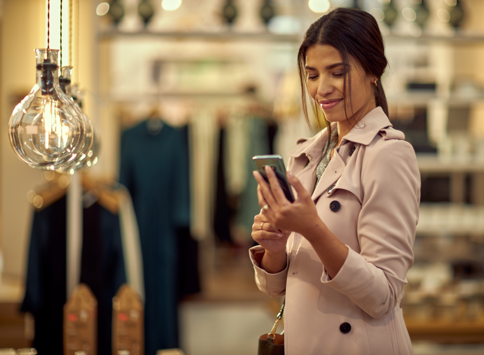 Mulher de casaco leve usando seu smartphone em uma loja de roupas com roupas e lâmpadas elegantes ao redor.
