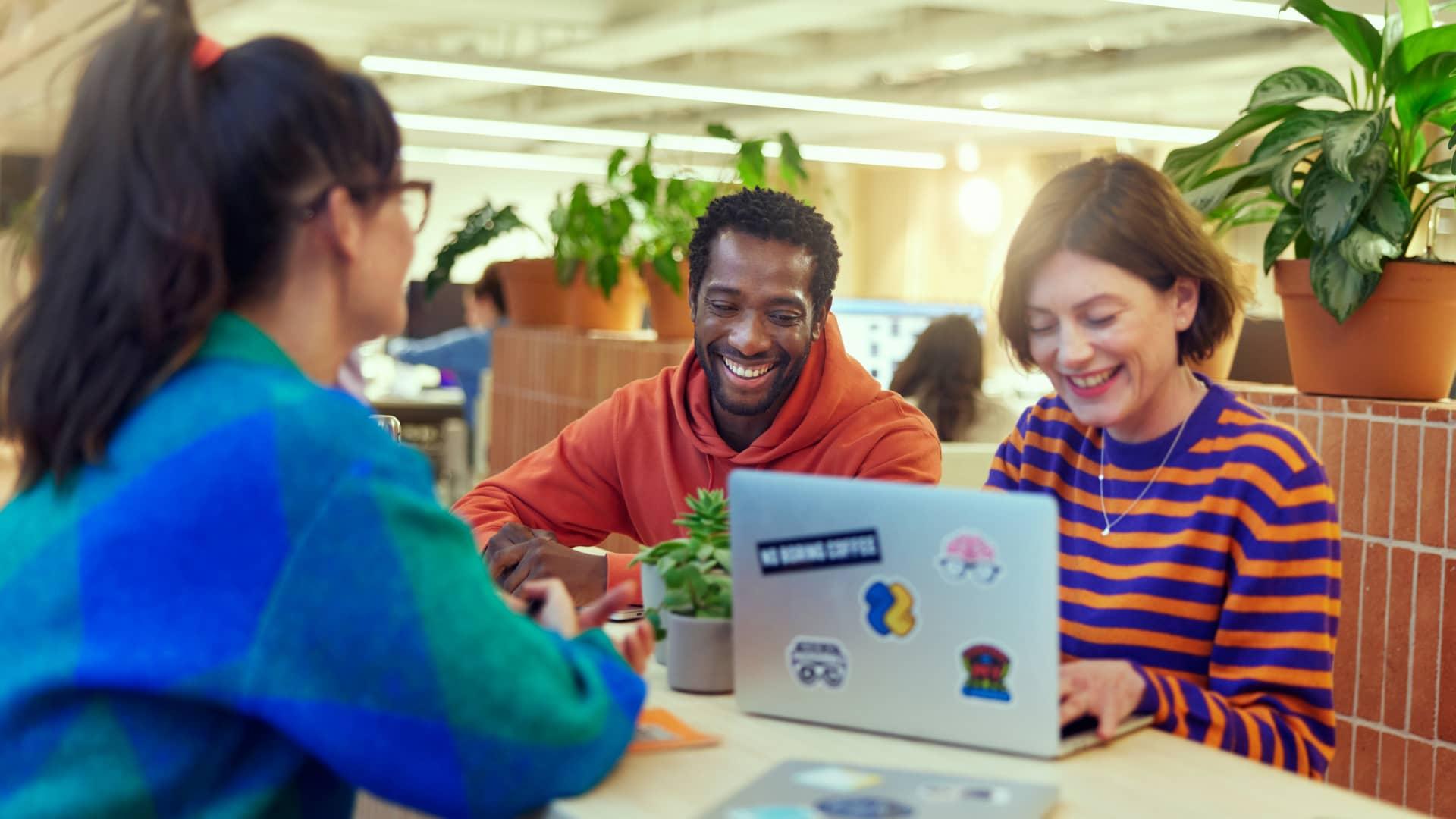 Drei Kollegen führen ein angenehmes Gespräch um einen Laptop in einer modernen Büroumgebung.